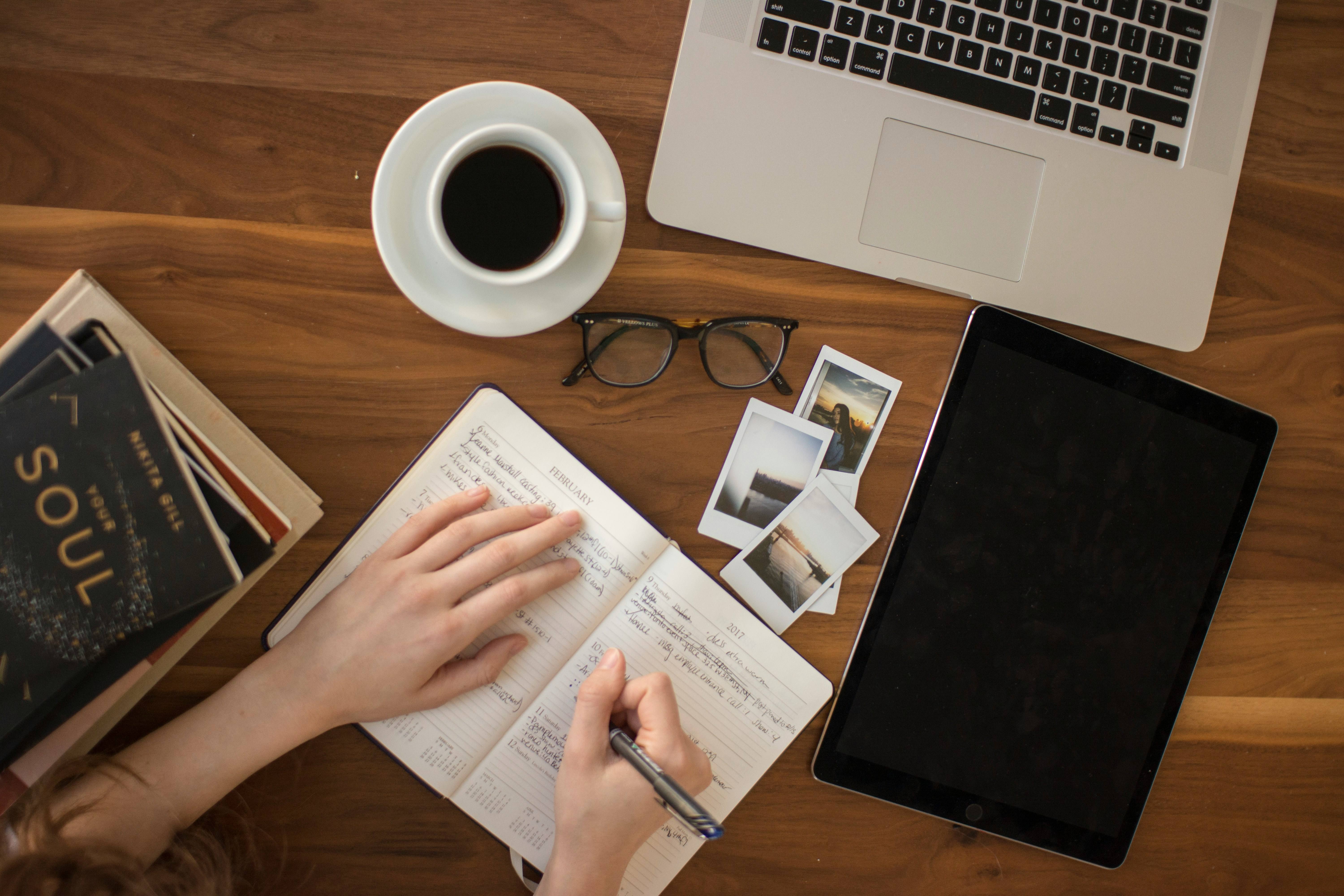 Overhead view of a person writing in a planner on a wooden desk with a laptop, tablet, and coffee.